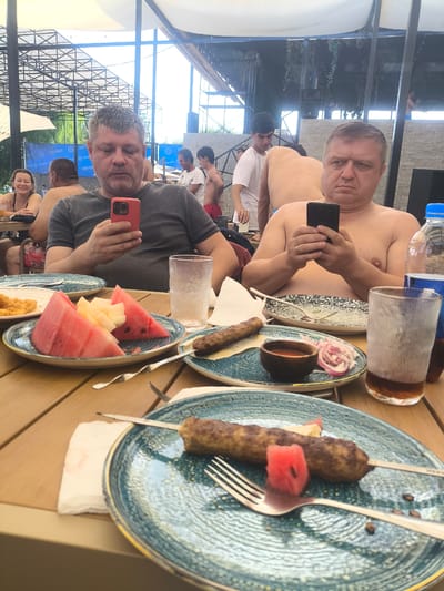 Two men are seated at a table outdoors, possibly at a restaurant or pool-side cafe in Vahdat, Tajikistan. Both men are looking at their mobile phones. The man on the left is wearing a grey t-shirt, and the man on the right is shirtless, showing visible sweat on his skin. In front of them are plates with food, including what appears to be grilled meat skewers, watermelon, and other fruits, as well as drinks. In the background, other people are visible, some of whom are also shirtless. The environment appears to be a covered outdoor area with a light-colored canopy overhead and some architectural structures in the background.