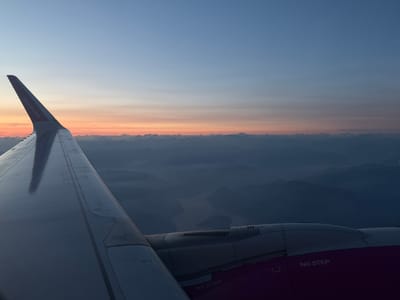 An aerial view shows an airplane wing and engine against a hazy sunset sky. Below the clouds, a body of water is visible, surrounded by hills. The "Wizzair.com" logo is visible on the wing. The phrase "NO STEP" is printed on the engine cowling. The specific location of Caravaggio, Italy, is not discernible from the image.