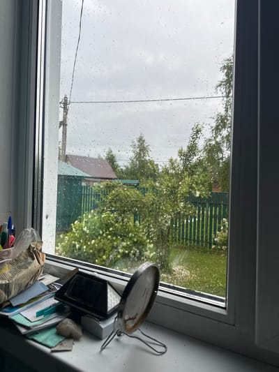The image depicts a view from a window in Красное, Russia, on a rainy day. Raindrops are visible on the glass, obscuring the outdoor scene to some extent. Outside, a green wooden fence encloses a yard with lush green grass and flowering bushes. Beyond the fence, a house with a brown roof and a green-roofed structure are partially visible, surrounded by trees. On the windowsill, various objects are scattered, including pens in a holder, crumpled paper, a small black electronic device with a solar panel, a clothespin, and a small rock. A round mirror with a metal stand is also present on the sill. The overall impression is one of a quiet, domestic scene observed during inclement weather.