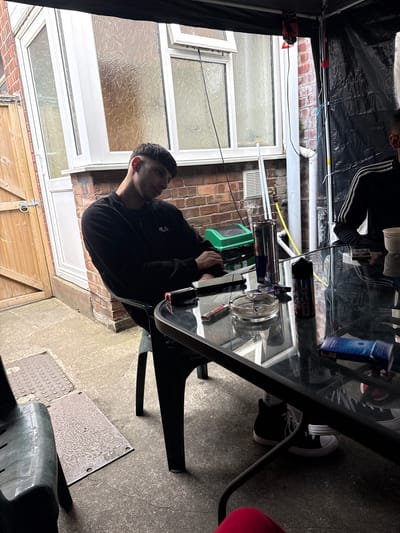 The image captures an outdoor patio or backyard scene at a residential property in Boston, United Kingdom. Two individuals are seated at a rectangular glass-top table with a black frame, situated under a dark tarp canopy.

On the left, a male with short, dark hair and a faded haircut is seated, looking downwards and slightly to the right. He is wearing a black long-sleeved jacket with a white "FILA" logo on the left chest. His right forearm rests on the table.

Partially visible on the right is another individual, whose upper arm and torso are clad in a black long-sleeved top featuring three white stripes, consistent with sportswear. Their lower legs and feet, adorned with black high-top sneakers with white soles, are visible beneath the table.

The glass-top table holds multiple items: a silver Red Bull energy drink can with blue accents, a large black bottle of vape liquid with a prominent "18+" label and cartoon graphics, a black smartphone, a circular glass ashtray containing cigarette butts and ash, at least two distinct lighters (one colorful, one red/black), a small white paper cup, a blue and white rectangular package (possibly cigarettes or rolling papers), and a smaller black package.

The background features a red brick wall, typical of British architecture, with a light-colored UPVC bay window on the left, fitted with patterned or obscure glass panes. A wooden garden door with a circular metal latch is visible to the far left. Various utility pipes and electrical cables run along the brick wall and support structure. A white ventilation grille is present on the lower section of the brick wall, and a yellow hose is coiled near the ground. The ground is concrete, showing wear and cracks, and features a textured metal access cover embedded flush with the surface, along with a separate flat, rectangular metal plate. Overhead, a black tarp provides cover, from which a red and black item and various cables are suspended. Two dark green plastic patio chairs are visible, occupied by the individuals.