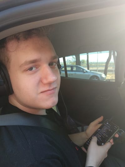 A young Caucasian male wearing headphones and a black t-shirt is seated in the back of a car, holding a smartphone. He is looking at the camera with a neutral expression. Outside the car window, a silver sedan is visible driving on a road, with trees and a fence in the background. The scene appears to be a sunny day. The image does not contain enough information to determine the specific location within the Municipal Unit of Vocha, Greece.