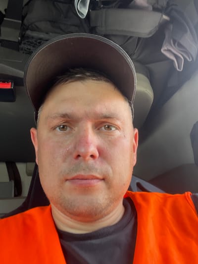The image is a close-up selfie of a Caucasian male wearing a dark baseball cap and an orange safety vest over a dark shirt. He has light brown eyes and stubble. The background appears to be the interior of a vehicle, possibly a truck cab, with dark fabric and upholstery visible. There are no visible landmarks or identifying features that would indicate a specific location in Jandelsbrunn, Germany. The lighting suggests an indoor or shaded environment.