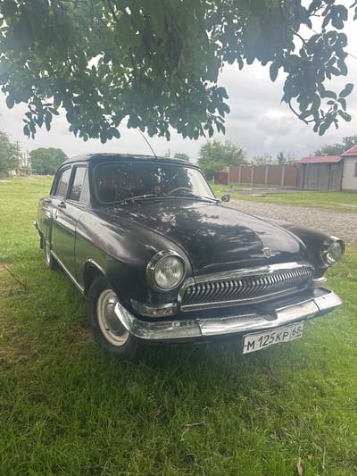 A black GAZ-21 Volga sedan is parked on a grassy lawn under a leafy tree. The car is facing slightly to the right of the camera, with its front end prominent. The license plate, which reads "M 125KP 68 RUS", is clearly visible on the front bumper. The background shows a fence, a building with a red roof, and more trees, suggesting a rural or suburban setting. The sky is overcast. The scene appears to be in Dzuarikau, Russia.
