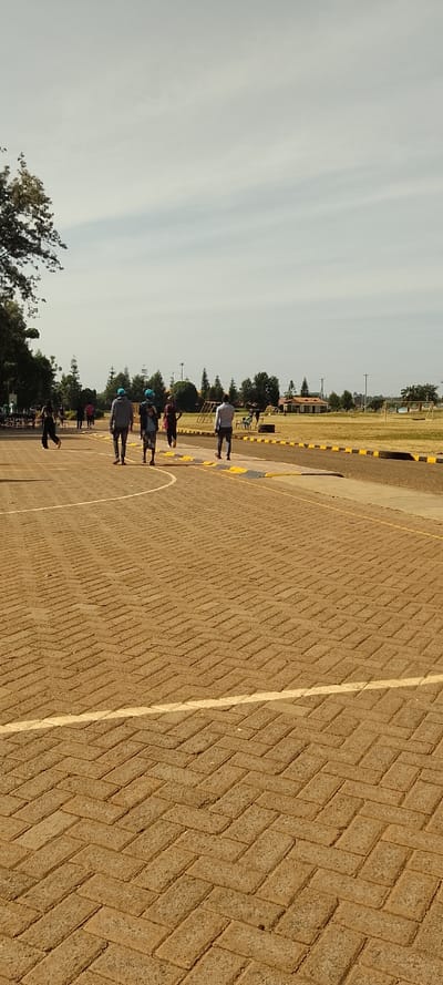 The image depicts an outdoor recreational area under a bright sky with scattered, wispy clouds, suggesting either morning or late afternoon light. In the foreground, there is a wide, light brown paved path constructed from interlocking pavers laid in a herringbone pattern. A prominent white curved line is painted across this path. To the right of the paved path, a darker strip of ground, possibly asphalt, is bordered by a series of yellow and black curb blocks or speed bumps.
The mid-ground and background are dominated by a large, open grassy field, which appears to be a sports ground, indicated by visible goalposts in the distance on the right side. The field is bordered by numerous trees, including some with dense green foliage and others resembling pine trees, particularly on the left side. A light-colored building with a reddish roof is situated on the far right edge of the field. On the left side of the field, nearer to the trees, there are structures that resemble children's playground equipment, such as swings and a slide, along with some tables and benches where a few individuals are gathered.
Several people are present in the scene, primarily in the mid-ground. A group of approximately five to six adults are walking casually along the paved path, generally moving away from the viewer. They are dressed in everyday attire; visible details include a person in a light blue cap and grey hoodie, and another with a blue cap and a backpack. On the far left of the image, near the trees, one person is depicted in motion, possibly running or playing. A few other figures are sparsely distributed across the distant grassy field. The overall atmosphere suggests a relaxed, open public space where people are engaged in casual outdoor activities. No text is visible in the image.