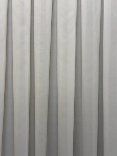 The image displays a close-up, top-down view of multiple horizontal layers of light-colored material, likely fabric, arranged in parallel folds or rolls. The dominant colors are white and various shades of light to medium gray, with darker gray shadows accentuating the spaces between the layers and creating a sense of depth. The material itself appears to have a slightly textured, possibly matte finish, and exhibits some minor variations in surface consistency, suggesting a textile or a non-woven fabric.
There are no people or other discernible objects present in the frame. The scene is static, depicting the arranged material without any ongoing activity. The lighting is even, consistent with an indoor setting, and does not indicate a specific time of day or weather conditions. There are no location-specific cues visible within the image itself, though the context provided specifies Kfar Saba, Israel. No visible text is present on the material or in the scene.