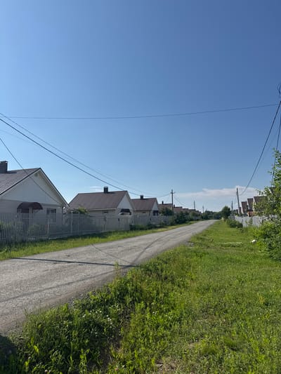The image depicts a residential street in the village of Yagul, Russia, on a clear, sunny day. The street is unpaved, appearing to be made of gravel, and curves gently into the distance. On either side of the street are houses with white fences. The houses are predominantly single-story with dark-colored roofs and light-colored siding. Electrical wires are strung across the sky, supported by utility poles. The foreground features lush green grass and some wild flowers. The overall impression is of a quiet, rural residential area.