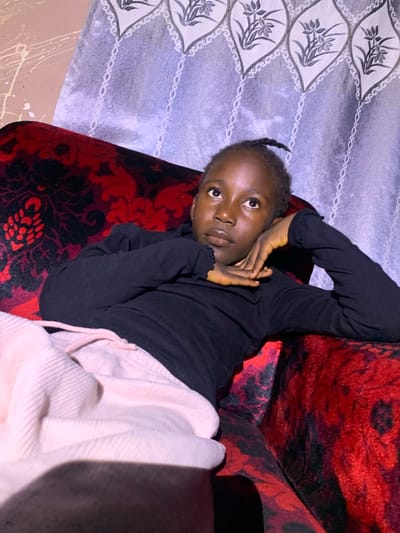 In an indoor setting, a young Black female is reclining on a red and black patterned sofa. She is wearing a long-sleeved black top and light pink corduroy pants. Her head is propped up on her hand, and she is looking upwards. The background features a patterned curtain that appears to be light blue and white. The overall lighting suggests an indoor environment, possibly at night due to the quality of the light. The image was taken in Russo, Nigeria.