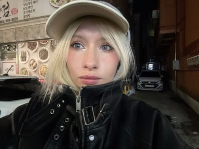 A blonde individual with blue eyes, wearing a beige baseball cap, a grey hooded garment, and a black leather-look jacket, stands in an urban outdoor setting. The person's gaze is directed towards the camera.

To the left, a building wall features a large sign for a restaurant named "남도밥상" (Namdo Bapsang) in Namyangju-si, South Korea. The sign displays various Korean dishes on circular plates and the phone number "0505-594-9929". The upper portion of a white vehicle is partially visible in front of the restaurant.

To the right and extending into the background, a narrow, dimly lit alleyway is visible. A white Chevrolet Spark hatchback is parked facing away from the viewer within this alleyway, with its license plate reading "372나 8921". The ground in the alley shows patches of snow or ice. Dark building walls line both sides of the alley.