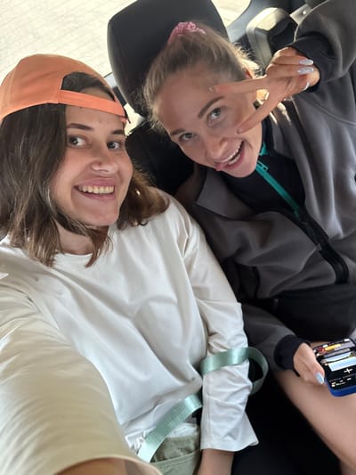 The image shows two young women in a vehicle, likely a car, as indicated by the background suggesting a window with light. The woman on the left is wearing an orange baseball cap and a white long-sleeved shirt, and she is smiling. The woman on the right is wearing a grey jacket and is making a peace sign with her fingers while sticking her tongue out. She has light-colored nail polish with small decorative dots. She is also holding a smartphone in her right hand, which displays an image of a landscape with a plus sign and text. The environment appears to be urban, possibly within the city of Zverosovkhoz, Russia, though this cannot be definitively confirmed from the image alone. The lighting suggests it is daytime.