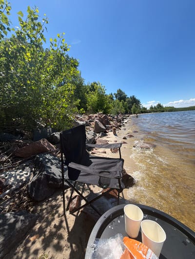 The image depicts a serene lakeside scene in Lebedivka, Ukraine, on a clear, sunny day. In the foreground, a black folding chair sits on a sandy beach, casting a shadow from the dappled sunlight filtering through nearby trees. A black tray is placed in front of the chair, holding two white paper cups and an orange packet. The beach is littered with a collection of rocks and driftwood, leading to the edge of a calm, wide body of water.

In the middle ground, the water laps gently against the shore, with small waves breaking. Further out, several people can be seen enjoying the water. The shoreline is lined with a dense growth of green trees and bushes, with a few larger rocks scattered along the edge. The sky above is a vibrant, cloudless blue, indicative of pleasant weather. The overall atmosphere is one of peaceful relaxation and enjoyment of nature.