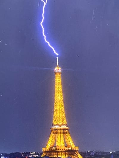 A vertical, full shot captures the Eiffel Tower in Paris, France, under a dark blue, clear night sky. A bright, jagged streak of blue lightning arcs from the top of the tower, reaching into the upper portion of the frame. The iconic iron lattice structure of the Eiffel Tower is illuminated in warm, golden light, highlighting its intricate details and tapering form. The base of the tower rests on a dark, blurred cityscape, suggesting the vastness of Paris at night. The image is slightly grainy and may have minor dust or scratches present. No people or animals are visible in the frame. The scene appears to be a moment captured during a lightning storm over the city.