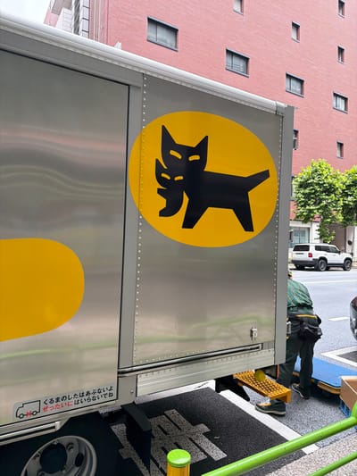 In Chuo, Japan, a silver delivery truck is parked on a city street. The truck features a prominent black logo of two stylized cats within a yellow oval on its side. Below the logo, Japanese text and a graphic indicating a truck with a red cross through it are visible, possibly a warning against unauthorized use. A person wearing a green and dark-colored uniform is seen near the truck's rear, standing on a yellow metal step. A white SUV is parked in the background on the street. The environment includes a red brick building with multiple windows and trees lining the sidewalk. The ground is paved with asphalt and there are green metal railings in the foreground.