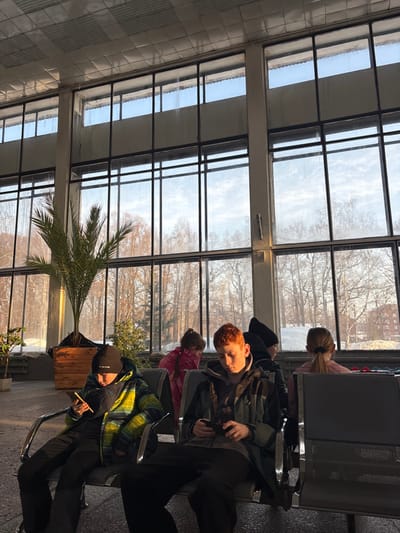 The scene depicts an indoor waiting area in the city of Дулесово, Russia, characterized by a wall of large, multi-pane windows looking out onto a winter landscape. Sunlight streams in from the exterior, casting shadows from the occupants and objects within the room.

Five individuals are present, seated on grey metal waiting chairs. In the foreground, a young male wearing a black beanie and a striped green, yellow, and black puffer jacket sits on the left, focused on a smartphone held in both hands. Adjacent to him, a young male with red hair, dressed in a dark jacket, also sits intently engaged with a smartphone. Behind these two, a young female wearing a pink jacket is partially visible, facing away from the camera. Further right, a person with a dark beanie or hood is partially obscured. On the far right, a young female with light brown hair, pulled into a ponytail, sits facing away and looking towards the right.

The interior features a tiled ceiling and a row of grey metal waiting chairs with perforated backs and seats. A large palm-like plant in a square wooden planter and a smaller potted plant are positioned near the windows on the left. The exterior view through the windows reveals bare deciduous trees and snow-covered ground. A multi-story brick building is discernible in the distance on the right side of the frame. The window panes exhibit evidence of condensation or streaking.