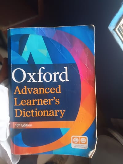 The image displays an Oxford Advanced Learner's Dictionary, 10th Edition, held by a person. The book's cover features a striking design with a dark blue background complemented by an abstract, multi-colored arc in shades of pink, orange, and red on the right, and geometric blue shapes including a prominent light blue 'A' on the left. The title "Oxford" is printed in large white letters, followed by "Advanced Learner's Dictionary" in orange text. "10th Edition" is shown in white on an orange band towards the bottom left. Two small white icons, depicting a book and a digital screen, are visible in the bottom right corner, indicating different available formats.

The dictionary exhibits visible wear, with several creases and scuff marks across the cover, particularly along the top edge and the right side, and a slightly worn bottom right corner. Part of a person's hand is visible on the left side, holding the book. The background is out of focus and appears dark, suggesting an indoor setting. A bright light source, likely daylight, illuminates the book from the right, creating reflections on its surface. The scene is set in Warri South, Nigeria.