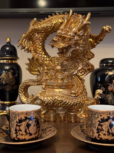 The image displays a decorative arrangement of Asian-inspired items. Dominating the foreground is a large, ornate, golden dragon sculpture, intricately detailed with scales and a fierce expression. Behind the dragon, and partially obscured, are several black porcelain items with gold floral and figural motifs. These include what appear to be a lidded jar or vase and at least two teacups and saucers. The teacups and saucers feature detailed gold patterns against a black background, with gold rims. The overall setting suggests an indoor display, possibly on a shelf or table, with a neutral background. The lighting is somewhat dim, highlighting the metallic sheen of the dragon and the decorative elements on the porcelain. There is no visible text in the image. The scene evokes a sense of opulence and traditional Asian aesthetics.