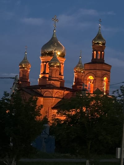 The image shows an Orthodox church in Novoteretsky, Russia, at dusk. The church is made of red brick and features several golden domes topped with crosses. The building is illuminated from within, casting a warm glow through arched windows. In the foreground, dark green trees partially obscure the view of the church. The sky is a deep blue with some clouds. Power lines are visible in the upper left and right corners of the image.