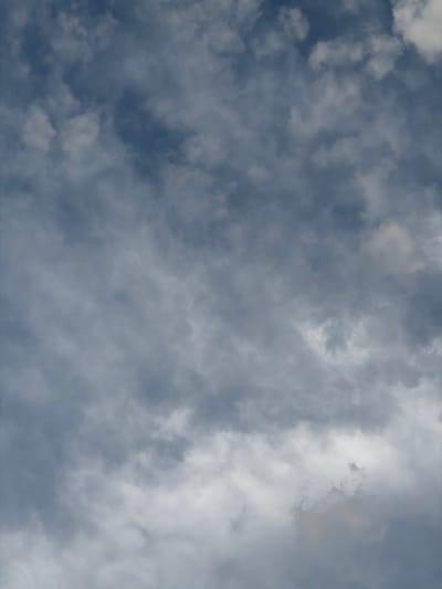 This is a medium shot, aerial view of a cloudy sky. The clouds are dense and varied in shade, ranging from a dark, stormy grey to bright white where the sun is beginning to break through. The overall impression is one of dynamic weather, possibly leading to or recovering from rain. The image is filled with the texture and movement of the clouds, with no discernible objects, people, or specific location cues other than that the scene is outdoors and under a vast expanse of sky. The lighting suggests it could be daytime, with the sun intermittently obscured by the thick cloud cover. There is no visible text in the image.