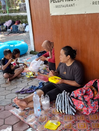The scene takes place in a location in the city of Kecamatan Tulung, Indonesia, at coordinates -7.592 latitude and 110.65 longitude. A woman wearing a black shirt and black pants sits on a colorful mat with a striped plastic bag beside her. She is eating with her right hand. Another woman wearing a hijab is sitting on the mat to her left, facing the same direction. A young boy and a young girl are sitting in front of them. There is a blue inflatable pool in the background, along with multiple backpacks and other objects. There is a sign on the wall of the building behind the women that reads "RESTAURANT." The scene is located in a public area adjacent to a building with a wooden facade. The surrounding environment appears to be a park or outdoor space with a paved area and vegetation. The group appears to be having a casual outdoor meal together.