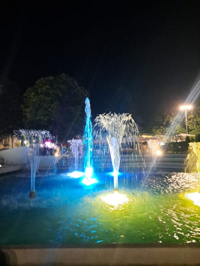 A night-time photograph captures a colorful water fountain display in the city of Popovo, Bulgaria. The fountain, illuminated by blue and yellow lights, shoots streams of water into the dark sky. In the background, trees and a lamppost are visible, as well as some structures suggesting an outdoor seating area or a building with lights. The water in the fountain reflects the vibrant colors of the lights, creating a dynamic and visually striking scene.