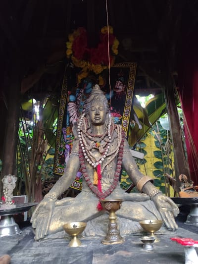 A medium shot shows a statue of a deity (Shiva) in the city of Bedulu, Indonesia, inside of what appears to be a small open-air shrine or temple. The gold-colored statue of Shiva is in a seated meditative pose, with hands resting on the knees. He is adorned with multiple strands of prayer beads around his neck. Behind the statue is a framed image of two figures, the top figure depicted as male with a mustache and the bottom figure blue. Behind the picture are green plants. Several small metal bowls and offering vessels are placed in front of the statue. The background shows a wooden structure and glimpses of the surrounding greenery, suggesting the temple is in a natural setting. Red and yellow garlands hang above the framed picture. Small statues are present on either side of the deity on stands.