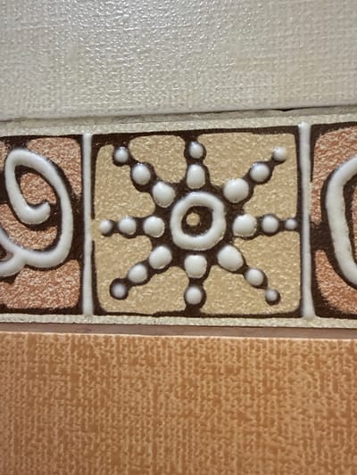 The image displays a close-up view of decorative tiles, likely from a building interior. The primary focus is on a central tile featuring a white, raised pattern resembling a stylized sun or starburst on a light beige background. This pattern is outlined in dark brown. To the left and right of this central tile are adjacent tiles with a textured, light brown and reddish-brown speckled pattern, also framed by dark brown and white borders. Above this row of tiles is a textured, off-white tile. Below, a solid, textured light orange tile is visible. The scene appears to be a static, architectural detail. There are no people or actions depicted. The lighting is even, suggesting an indoor environment. The location is Taldybulak, Kazakhstan, but the specific context within the city is not discernible from the image alone.