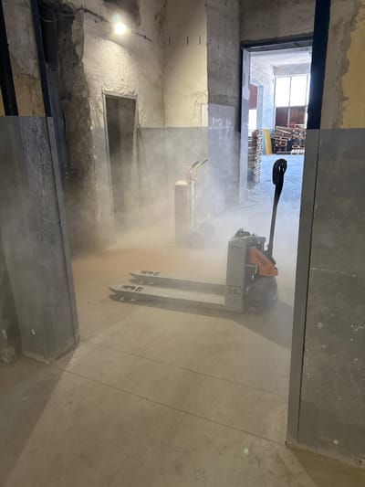 The image shows an interior scene, likely a warehouse or industrial space, in Djerman, Bulgaria. A pallet jack, possibly electric, with the brand "STILL" visible, is the central object. It is positioned on a concrete floor, partially obscured by a cloud of dust or mist. To the left, there is a doorway with a dark opening, and beyond that, a rough concrete wall. In the background, through another doorway, stacks of wooden pallets are visible, along with daylight entering the space. A hand truck is also present in the background. The walls appear unfinished and gritty, consistent with a construction or industrial environment.