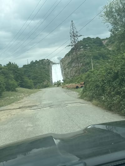 The image shows a view from a vehicle traveling on a paved road towards a large waterfall cascading down a steep, rocky cliff face. The waterfall is the focal point, with its white water contrasting sharply against the gray rock. Tall, dark green trees and lush vegetation cover the surrounding hillsides. In the background, a massive, skeletal metal pylon, part of a power line system, stands on a higher hill to the right of the waterfall, with several thick power lines stretching across the overcast sky. On the left side of the road, closer to the viewer, a section of the embankment is covered in grass and smaller shrubs. Several large, rusty pipes are visible on the ground to the right of the road, partially obscured by dense green foliage. The sky is cloudy and gray, suggesting an overcast or possibly rainy day. The scene appears to be located in a mountainous or hilly region, and based on the prompt's information, it is identified as Zhinvala, Georgia. The perspective is from inside a vehicle, with the hood of the car visible in the foreground.