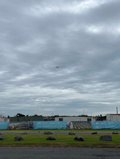 The image, taken during daytime under an overcast sky, is dominated by a large expanse of cloud cover. A small airplane is visible in the upper-left quadrant, flying across the cloudy backdrop.

The lower portion of the image depicts a ground-level scene. A strip of green grass runs horizontally, dotted with several dark, irregularly shaped rocks. To the immediate right of the grass is a paved surface, likely a road or path. Beyond this, a construction site is visible, characterized by several buildings and a long stretch of blue construction fencing.

Visible text on the blue fencing provides location cues. Logos for "BMD" and "Northshore Brisbane" are clearly discernable. Phrases such as "A Place of Connection" and "Sustainable Design" are also printed on the banners attached to the fence. These details, combined with the Hamilton, Australia context, suggest the image was taken near the Northshore Brisbane development. The overall impression is of an urban or industrial area with ongoing development, viewed on a cloudy but bright day.