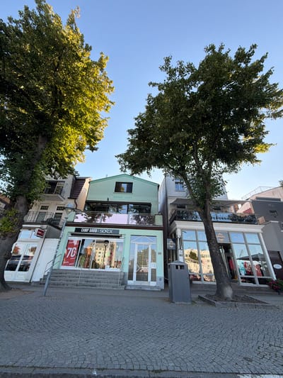The image shows a street scene with buildings and trees in Rostock, Germany. The foreground is dominated by a cobblestone street. Several trees with green foliage are present, with one large tree on the left and another on the right.
The buildings appear to be commercial establishments. The building in the center is painted light green and has large glass windows. A sign above the windows reads "CAMP DAVID SOEX". A "70% FINAL SALE" sign is visible in the window of this building. The building to the right of it is grey and also has large glass windows, with a sign visible that says "SARAZUL". Numbered signs "98" and "99" are visible on the facades of the buildings to the left and right respectively, indicating shop numbers.
In the background, more buildings are visible, including one with a balcony. The sky is clear and blue. The overall impression is a sunny day in a European city, likely a commercial area.