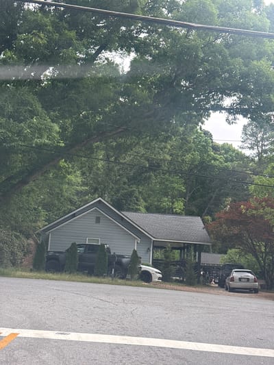 The image depicts a residential street in Mountain Park, Georgia, United States. A light blue, single-story house with a gray roof and a triangular front gable faces the road. Multiple vehicles are parked in the driveway and along the side of the road next to the house, including white, black and silver cars. The street is asphalt with a white line marking the edge. Abundant green trees and vegetation surround the house and line the street. Power lines are visible above the house and trees.