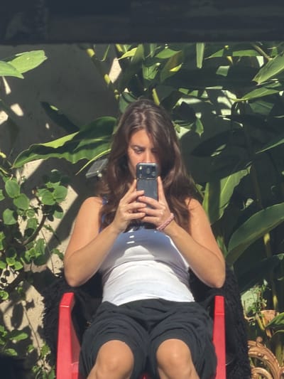 A woman is outdoors, holding a smartphone with both hands as if taking a picture. She has long brown hair and is wearing a white top and black shorts. She is sitting on a red object which rests on a black, fuzzy surface. The background includes various green plants and a light-colored concrete surface. The specific location in Vicente López, Argentina, cannot be determined from the provided image.