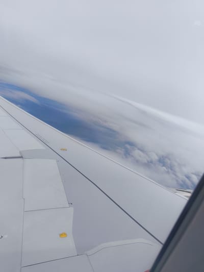An aerial view from an airplane window shows the wing of the aircraft angled across the frame. The wing is primarily white, with visible panel lines and a thin, dark gray line running diagonally from the upper right to the lower left. A small, yellow object, possibly a cover or indicator, is situated on the wing's surface. Below the wing, a landscape of blue sky and white clouds is visible, indicating the aircraft is in flight at a high altitude. The scene is dominated by the wing and the cloud-filled sky, suggesting an overcast or partly cloudy day. There are no discernible features that would indicate the specific location within Lam Son, Vietnam.