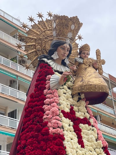 A large, ornate religious float featuring two statues is prominently displayed in a street in Paiporta, Spain. The central figure is a female statue, wearing a golden crown adorned with silver floral motifs and a diamond-shaped jewel on the forehead, large ornate white earrings, and a ruffled white collar. Her left hand holds a small green object. Behind her head is an elaborate golden sunburst or halo, featuring numerous radiating points topped with stars. To her right stands a smaller child statue, also crowned in gold and wearing a ruffled white collar, holding a decorated golden cross with both hands.

The lower portion of the female statue's robe is densely covered with fresh flowers arranged in horizontal bands and patterns. The colors include deep red, light pink, and white. A distinct red cross pattern is visible against a field of white flowers on the lower right side of the float.

In the background, a multi-story residential building with light-colored facades and balconies is visible. The balconies feature metal railings and light blue-green awnings or trim. Air conditioning units are attached to some of the balcony walls. The sky above is overcast and uniformly light grey.