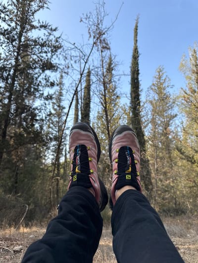 The scene presents a low-angle, first-person view of an individual's lower legs and feet against a natural backdrop. The subject wears black trousers and two matching Salomon brand trail running shoes, which are predominantly pink-salmon with black soles and a black Quicklace lacing system featuring yellow and blue accents. The feet are elevated, indicating a seated or reclined posture. The immediate ground beneath the feet consists of dry, light-colored soil with scattered natural debris. The background is dominated by a dense forest, featuring numerous evergreen trees, including several notably tall and slender cypress-like trees, interspersed with some deciduous trees exhibiting bare branches. A clear, bright blue sky is visible above the tree line. This forested environment is consistent with natural parklands or wooded areas within or immediately surrounding the city of Modi'in-Maccabim-Re'ut, Israel.