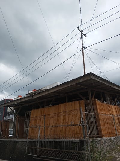 An outdoor scene in Cibadak, Indonesia, depicts a partial view of an open-sided storage or workshop structure under a heavily overcast, grey sky. A tall, dark utility pole topped with insulators and numerous black electrical or communication wires crisscrosses the upper frame, extending from the pole in multiple directions. The structure below features a corrugated metal roof supported by dark wooden beams and posts. Inside, large quantities of light brown wooden planks are stacked vertically, filling much of the visible interior. A dark, rusted metal fence with a diamond pattern base and vertical bars forms a gate in the immediate foreground, separating the viewer from the structure. Below the fence and structure is a stone wall foundation with minimal green vegetation and a narrow, dark ground channel. In the background, to the left of the main structure, parts of other buildings are visible, including a dark building facade with white lettering and a white-walled building featuring three orange cylindrical water tanks on its roof. No individuals or active events are present.
