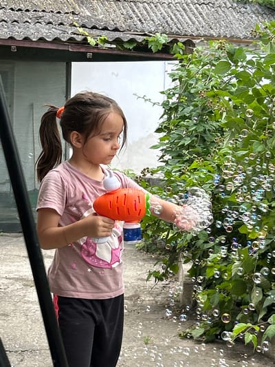 A young girl in Prokhladny, Russia, is pictured outside on a cloudy day, holding a toy bubble gun shaped like a carrot. She is wearing a pink t-shirt with a unicorn design and black pants. The bubble gun is actively shooting bubbles, which fill the air around her and the surrounding green foliage. In the background, there's a textured grey wall and a corrugated roof. The girl appears engaged and happy as she plays with the bubbles.