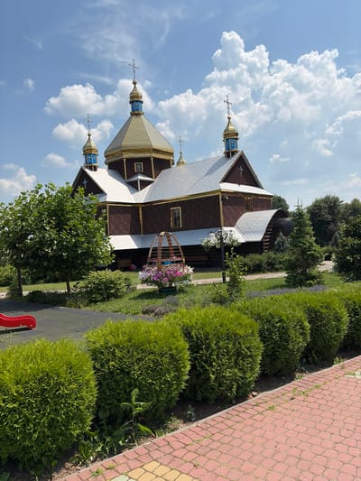 A wooden church with a golden dome and blue accents stands under a cloudy sky. In the foreground, neatly trimmed green bushes line a brick pathway. To the left, a portion of a red playground slide is visible. Lush green trees and well-maintained landscaping surround the church. The scene appears to be in the city of Triitsia, Ukraine, during daylight hours.