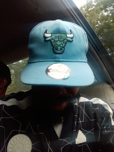 The image shows a person wearing a light blue baseball cap with a green and black Chicago Bulls logo. The person is inside a vehicle, likely a car, with trees visible through the rear window. The person is also wearing a black and white patterned shirt. The scene is captured from a frontal perspective.