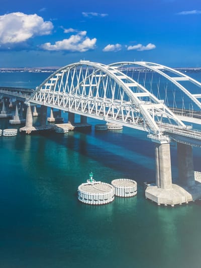 The image depicts the Kerch Strait Bridge, a massive infrastructure project connecting Russia's Taman Peninsula to the Kerch Peninsula of Crimea. The bridge is an impressive feat of engineering, featuring multiple spans and arches. It crosses a body of water that appears to be calm and a deep blue-green color. In the distance, a shoreline with some structures can be seen under a bright blue sky with scattered white clouds. The bridge itself is a prominent white structure, with its intricate metal framework visible. Several concrete piers support the bridge above the water. In the foreground, circular structures are visible in the water, likely navigational aids or support elements for the bridge. One of these circular structures has a green beacon on top. The scene suggests a clear day with good visibility, highlighting the grandeur and scale of the bridge against the natural backdrop of the sea and sky. The city of Минзаг is not depicted in this image.