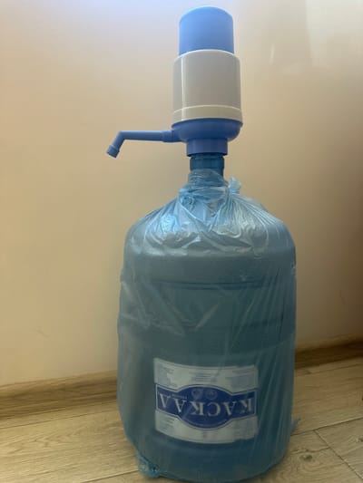 A blue and white water pump is attached to a large, clear plastic water jug. The jug is covered in a crinkled blue plastic bag, and sits on a light brown wooden floor against a plain, light-colored wall. The scene is indoors, and appears to be a domestic or office setting. There are no people or animals visible in the image. The location is Orlivka, Ukraine.