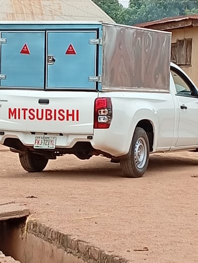A white Mitsubishi pickup truck is parked outdoors, viewed from its rear three-quarter angle. The truck features a custom-built cargo box in its bed, which is light blue at the back with two doors, and metallic silver on its visible side. Each blue door has a red triangular warning sign with a white symbol, possibly an 'A', inscribed within it. The tailgate of the truck clearly displays the word "MITSUBISHI" in prominent red lettering.
A Nigerian license plate, partially visible, reads "FXJ-721YL". Above the numbers, "BENUE STATE" is faintly discernible, consistent with the provided location context of Gboko. The vehicle's rear wheel, a standard steel rim, is visible, resting on unpaved, reddish-brown ground.
In the immediate foreground, a concrete and stone open drainage channel or gutter runs across the image. The background is somewhat blurred, showing simple structures, possibly houses with corrugated roofs, and green foliage under what appears to be an overcast sky or during the late afternoon, given the soft lighting. No specific activity is depicted; the truck appears stationary. No people are visible. The scene suggests a setting in a rural or semi-urban environment in Gboko, Nigeria.