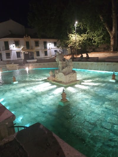 The image depicts a nighttime view of the Fuente del Rey in Priego de Córdoba, Spain. A large, ornate fountain with turquoise-colored water is the central focus. The fountain features a sculpture at its center and several smaller decorative elements. The water is illuminated from below, creating a vibrant glow. In the background, a white building with a colonial architectural style is visible, with windows and balconies. To the right of the fountain, trees with lush foliage are present, with a street lamp casting light on the scene. Several individuals are seated on a stone ledge in the background, observing the fountain. The overall atmosphere is serene and illuminated.