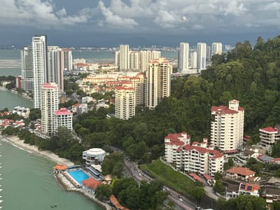 This is an aerial view of a coastal city, likely George Town, Malaysia, on a cloudy day. The foreground features a turquoise swimming pool with surrounding buildings with red-tiled roofs, situated near a sandy beach and the ocean. A road with moderate traffic runs alongside the beach, bordered by lush green trees and more residential buildings. In the mid-ground, several tall, modern apartment buildings dominate the skyline, interspersed with more traditional houses. Further in the distance, a sprawling city extends towards a hazy horizon, with mountains visible beyond. The sky is filled with dramatic, dark clouds, suggesting an impending storm. There are no people clearly visible in the image.