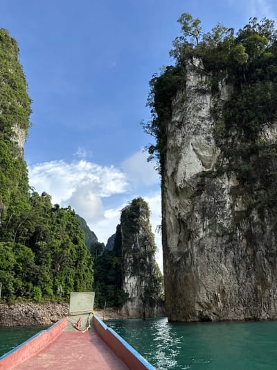 This media file shows a boat on a body of water, possibly a lake or river, passing between two large, steep, tree-covered rocky cliffs. The water is a vibrant turquoise color, and the sky is a clear blue with scattered white clouds. The sun appears to be shining brightly, suggesting it is daytime. The perspective is from a boat, with the bow of the boat visible in the foreground on the right side. The boat is red and blue. The cliffs are covered in lush green vegetation, and the rock faces show layers and textures. In the distance, more green hills and rock formations can be seen under the bright sky. The scene conveys a sense of natural beauty and tranquility, with the boat appearing to navigate through a scenic passage in the rocky landscape. There is no visible text.