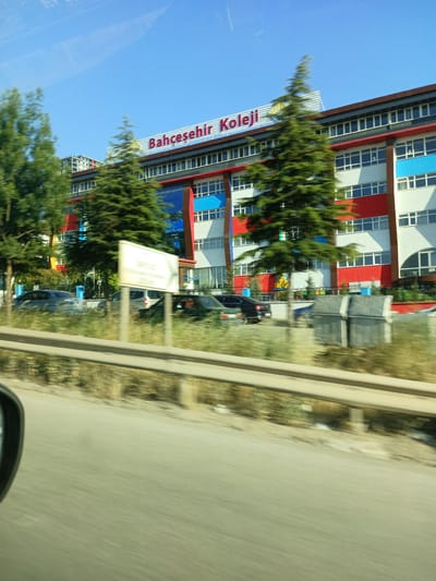 A low-angle, exterior shot captures the "Bahçeşehir Koleji" school building in Yahşihan, Turkey, under a clear blue sky. The building features a modern architectural style with a multi-colored facade of white, red, and blue panels, accented by large windows. Several mature evergreen trees stand in front of the structure, their foliage partially obscuring the building. A sign with text is visible in the foreground, positioned on a pole. Multiple cars are parked or driving in front of the school, and two large, grey trash receptacles are located near the building's entrance. The scene is captured from a moving vehicle, indicated by the motion blur in the foreground and on the road surface. The overall impression is one of a bright, sunny day in an urban or suburban environment.