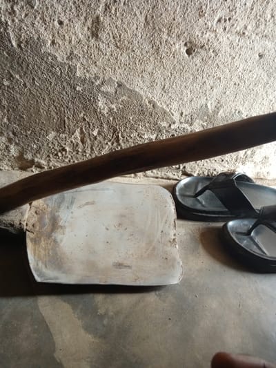 The image shows a close-up, eye-level view of several objects on a concrete floor in front of a rough, textured, light-colored wall. On the left, a light-colored, rectangular metal object with a slightly curved edge is visible. A dark, cylindrical wooden pole runs horizontally across the middle of the image, resting on or in front of the metal object and the wall. To the right of the metal object and partially under the wooden pole are two black flip-flops. The wall behind the objects is uneven and appears to be made of cement or plaster, with visible texture and some darker patches. The concrete floor in the foreground is plain and shows some lighter and darker areas, possibly from moisture or dirt. The lighting suggests an indoor or shaded environment. The scene is likely located in Gindin Dutse, Nigeria, given the context of the request.