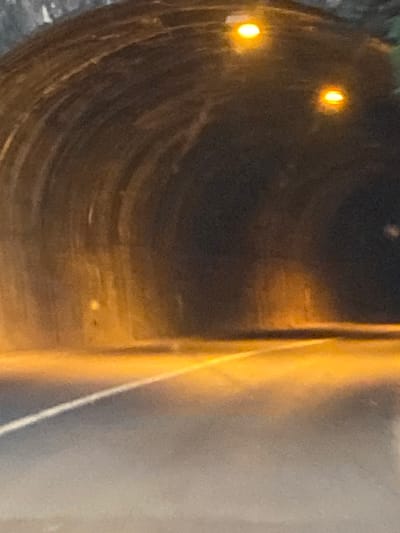 The image depicts the interior of a tunnel, likely on a road in Meljine, Montenegro. The tunnel walls are dark and curved, constructed from rough, textured material, possibly stone or concrete. Two bright, circular yellow lights are visible on the ceiling of the tunnel, illuminating the darkness. The road surface is visible in the foreground, appearing dark and wet, with a white line marking its center. The overall lighting is dim and atmospheric, with a strong orange-yellow hue cast by the tunnel lights. The scene suggests a typical road tunnel environment, possibly at night or during overcast conditions, with the limited visibility characteristic of such a passage.