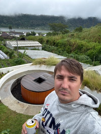 A man in a gray hooded sweatshirt takes a selfie. He holds a can of Bintang beer. He is standing near a round wooden structure with a square cutout in the center, surrounded by a concrete base. Lush green vegetation and greenhouses extend behind him. Fog-shrouded mountains are visible in the distance. The overall scene suggests a high-altitude, possibly tropical location.
