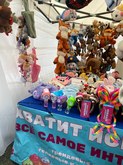 This image shows a close-up view of a market stall filled with an assortment of plush toys and novelty items. The stall appears to be set up outdoors, possibly under a tent or awning, indicated by the white fabric in the background and the metal poles. Various stuffed animals, including cartoon characters and generic animal shapes, are displayed, many hanging from a rack. Among the plush toys, there are also bubble wands and other small plastic toys.
A prominent element in the foreground is a large, light blue banner with bold, white and pink lettering. Partially visible text on the banner reads "ВСЕ САМОЕ Инт" which likely translates to "All the Best/Most Important". Another visible phrase on the banner appears to be "Гелиевые трендовые", potentially meaning "Helium Trendy". There are also two pink boxes with text that reads "MERMAID BUBBLE STICK". The presence of these items suggests a fun, carnival-like atmosphere, possibly geared towards children or impulse purchases. The lighting suggests it is daytime, with no strong indicators of specific weather conditions. The overall impression is one of a busy, colorful market stall offering a variety of playful merchandise.