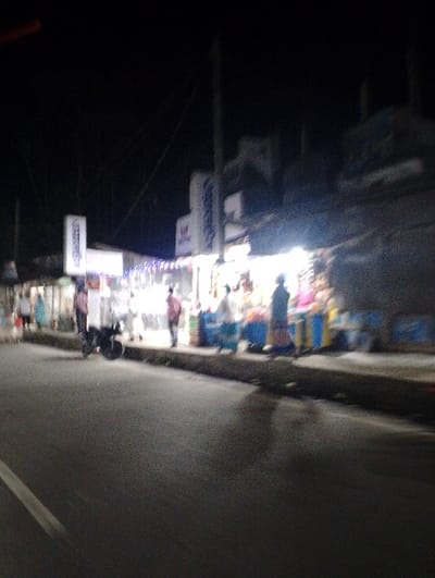 The image shows a nighttime street scene in Pangasia, Bangladesh, located at the coordinates 22.696 latitude and 90.598 longitude. The street is paved and appears to be relatively empty, with a few people walking along the sidewalk. There are several buildings lining the street, some of which are brightly lit.  One building has a sign that says "W.T. Super." Several people are standing in front of the buildings, likely waiting or shopping. A motorbike is parked on the street, facing towards the right. Some trees are visible to the left of the image. The scene is illuminated by artificial light from the buildings and streetlights. The overall image quality is grainy and blurry.